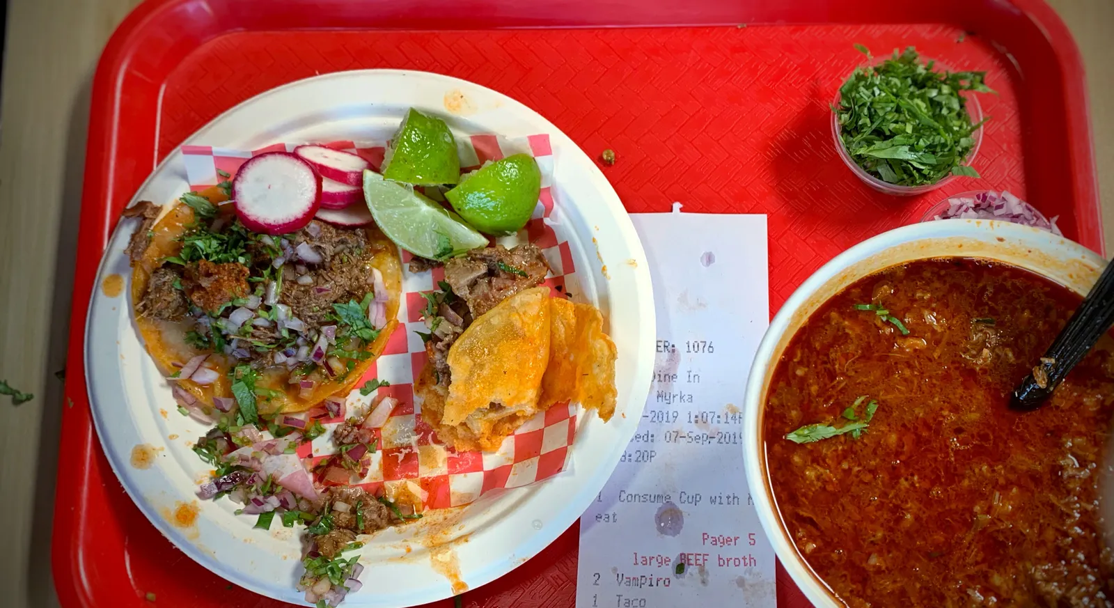 Birria tacos with cilantro, onion, radish and lime on a paper plate, beside a bowl of red birria consomme for dipping