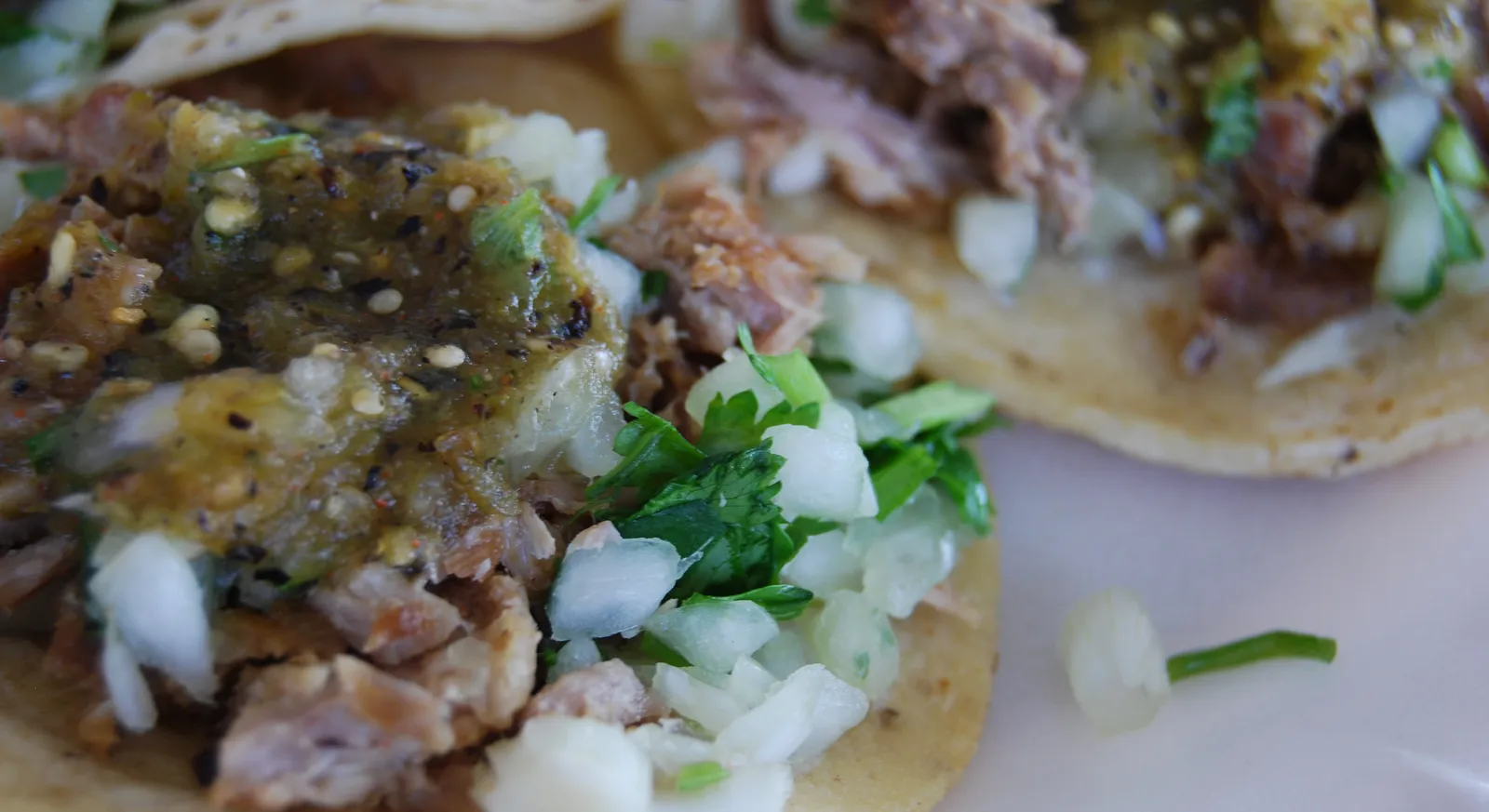 Close-up of carnitas tacos on corn tortillas topped with salsa verde, chopped white onion and cilantro