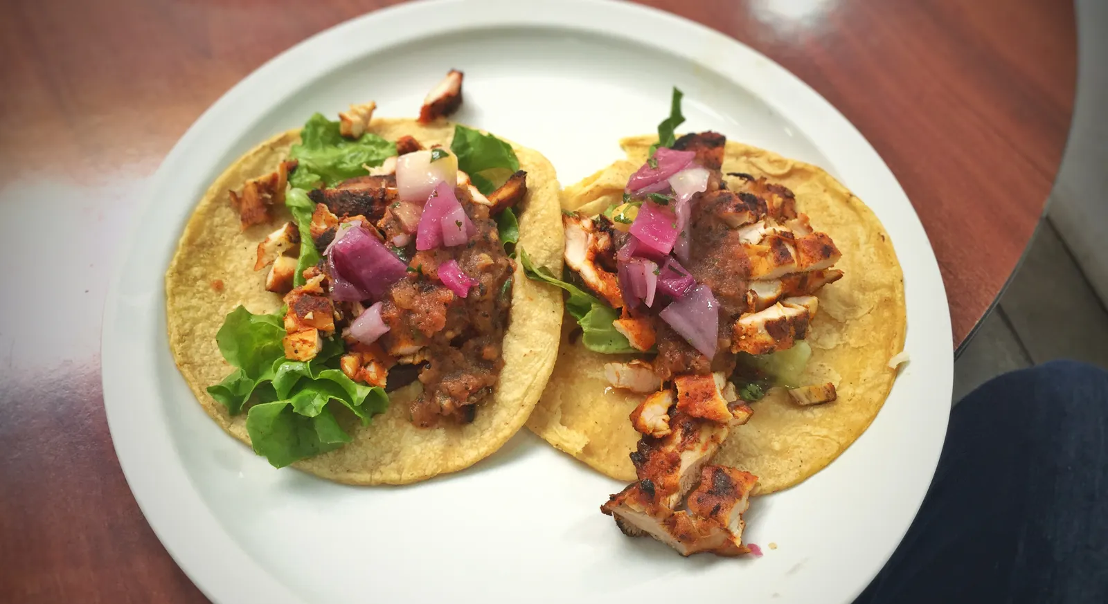 Two pollo asado chicken tacos on corn tortillas with lettuce, pickled red onion and fire-roasted tomato sauce
