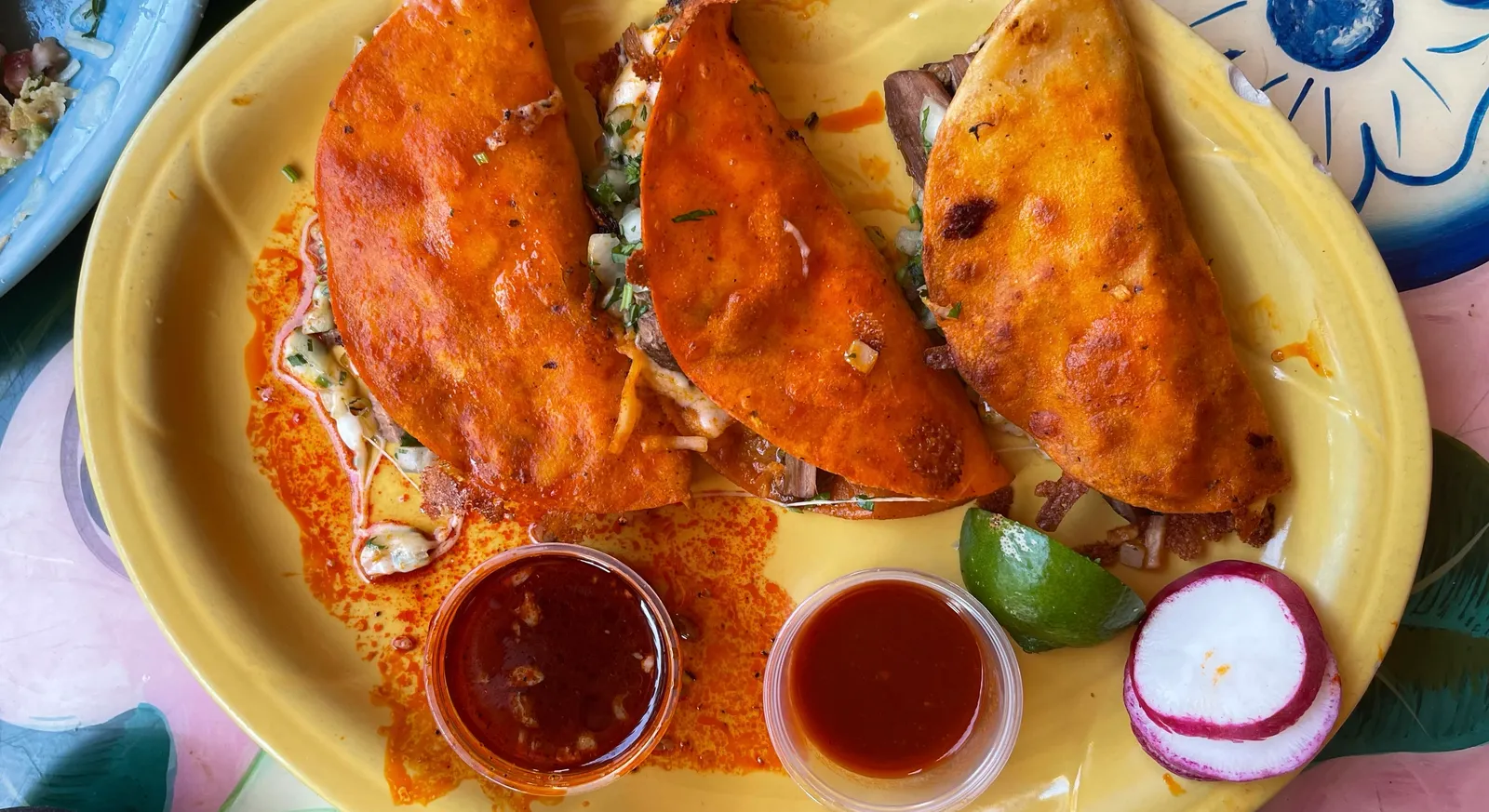 Three red-griddled quesabirria tacos with two salsas, lime and sliced radish on a yellow plate at Taqueria La Hacienda in Sonoma, California