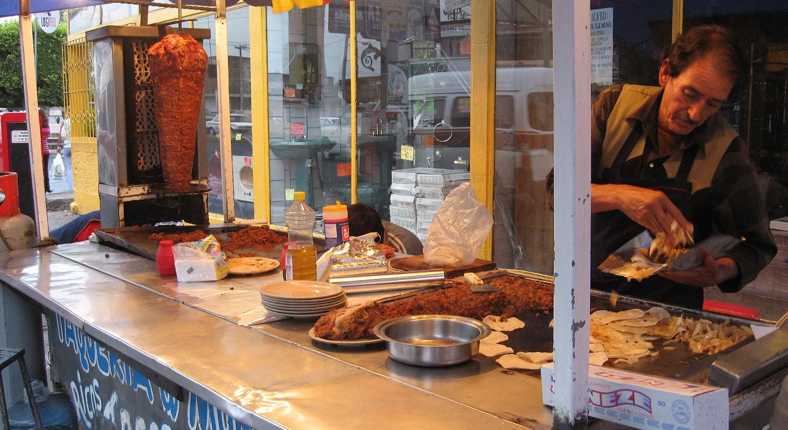 A taquero preparing tacos at a taqueria counter beside the al pastor spit