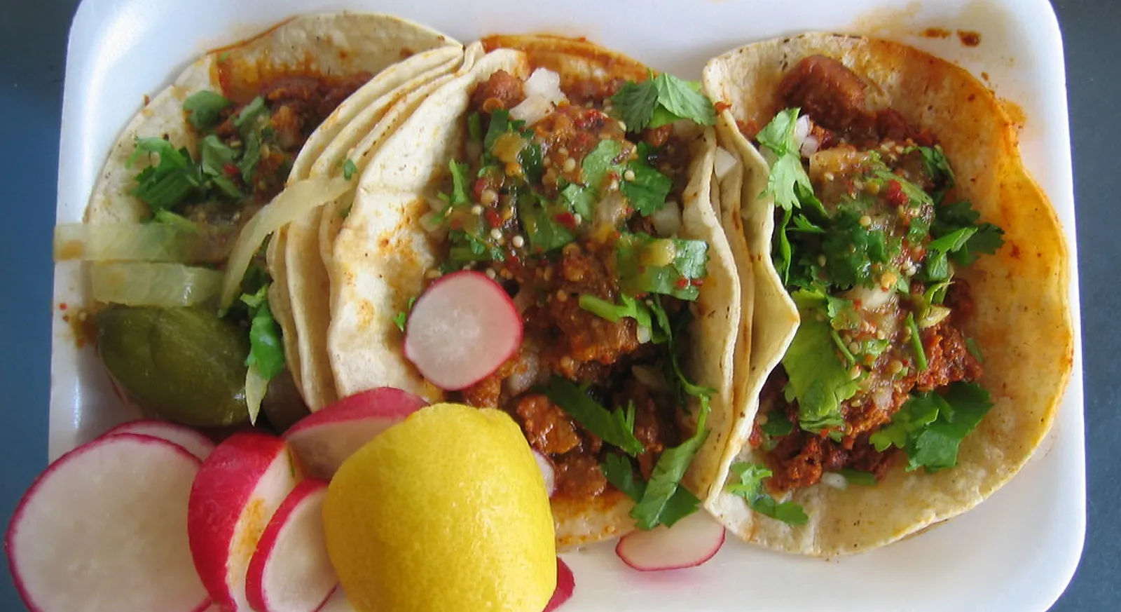 Three tacos with cilantro, onion, radish and lemon on a plate